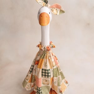 May include: A white goose figurine featuring an orange beak and orange webbed feet. The goose wears a ruffled dress and a matching bow on its head, both made of fabric with a patchwork pattern of pumpkins, green gingham, and floral motifs.