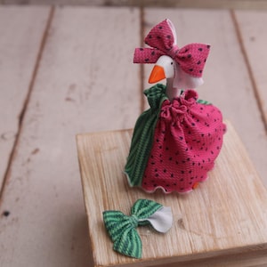 Puede incluir: Una figura caprichosa de ganso con un vestido rosa con lunares negros y un lazo a juego. El ganso tiene un pico naranja y un lazo verde y blanco. La figura está sobre un bloque de madera.