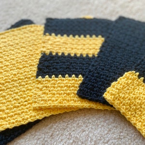 May include: Four crocheted dishcloths in a yellow and black color scheme. The dishcloths are made with a textured stitch and have a square shape.