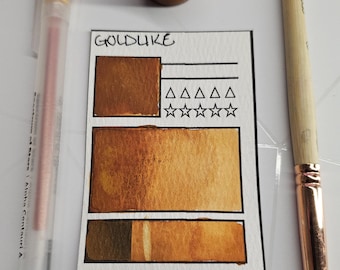 May include: A watercolour paint swatch card labelled "GOLDDUKE" with shades of gold and brown. A small jar of paint, a pen, and a paintbrush are also present. The card includes a rating system and the text "G: y/n".