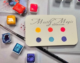 Könnte beinhalten: Eine Karte mit dem Text "Mostly Magic Watercolors" und Farbmuster von gelber, roter, blauer und violetter Aquarellfarbe.