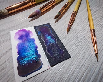 May include: Two swatches of iridescent blue and purple ink on white and black paper. The ink has a shimmery finish. Several paintbrushes are visible in the background.