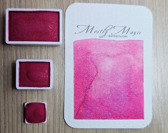 May include: Three watercolour paint pans in white plastic containers. The pans are filled with a shimmery, deep pink watercolour paint. The largest pan is labelled "Mostly Magic Watercolours".