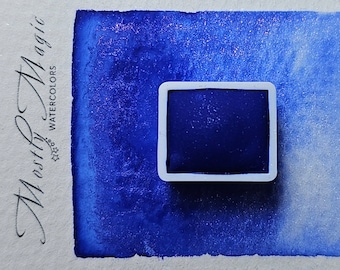 May include: A single watercolour paint pan in a white plastic tray. The pan is filled with a dark blue watercolour paint. The paint is wet and has a shiny, metallic finish. The pan is labelled "M.isty Magic Watercolours".