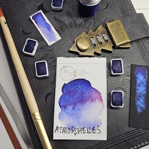 May include: An assortment of art supplies, including watercolor paints, a brush, and a notebook. The watercolor paints are in various forms, including small square pans and a jar. A small card with the word "ATMOSPHERES" is also present.