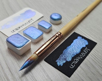 May include: A set of four watercolor paint pans in a light blue iridescent color with the word "ULTRAVIOLET" written on the label. The paint pans are on a wooden surface with a paintbrush and a small sample of the paint.