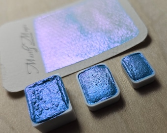 May include: Three watercolor paint pans with a shimmery, iridescent finish. The pans are in different sizes and are arranged on a wooden surface. The paint is a deep blue with a purple sheen.