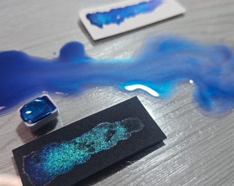 May include: A close-up of blue watercolor paints, including a small square of paint in a white container, a puddle of paint, and swatches on white and black paper. The paints have a shimmering, metallic finish.