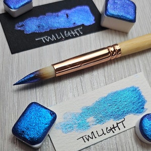 May include: Two pans of blue iridescent watercolor paint with the word "TWILIGHT" written on each pan. A paintbrush is laying on a wooden surface.