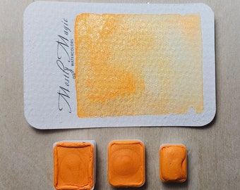 May include: Three watercolour paint pans in a light orange colour. The pans are labelled "Merry Magic Watercolours".