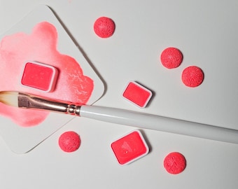 May include: A white surface with a pink watercolor palette, a paintbrush, and several pink round objects. The palette has a wet pink watercolor wash on it.