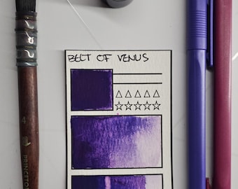 May include: A collection of art supplies including a watercolour brush, a small jar with a silver lid, and two markers. A card displays the text "BELT OF VENUS" with purple watercolour swatches and a rating system. The markers are purple and magenta.