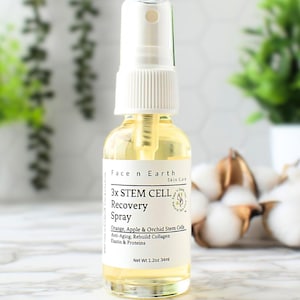 May include: A clear glass bottle with a white spray top and a white label. The label says "3x STEM CELL Recovery Spray" and lists ingredients including orange, apple, and orchid stem cells. The bottle is sitting on a white marble surface.