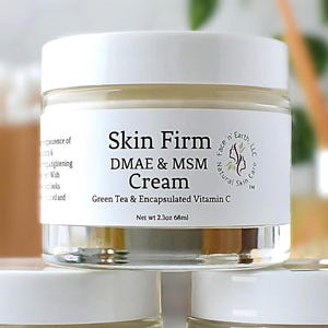 Könnte beinhalten: Drei klare Glastiegel mit Skin Firm DMAE & MSM Creme, weißen Deckeln und Etiketten, die jeweils 68 ml Produkt enthalten. Ein kleiner Probentiegel mit weißem Deckel und der Aufschrift Skin Firm DMAE Cream Sample steht vorne in der Mitte.