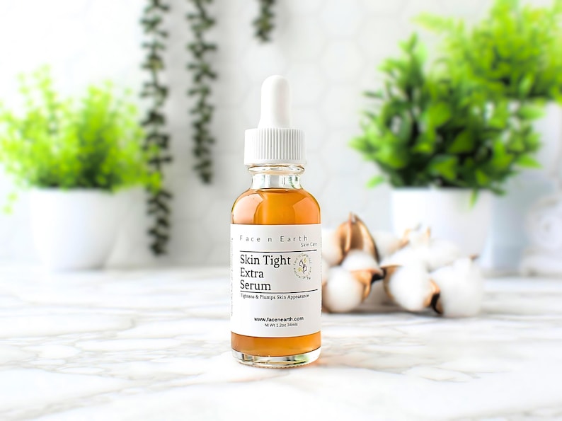 May include: A clear glass bottle with a white dropper top filled with a light brown liquid. The bottle label reads "Face n Earth Skin Care" and "Skin Tight Extra Serum" with the tagline "Tightens & Plumps Skin Appearance". The bottle is sitting on a white marble surface with green plants and white cotton balls in the background.