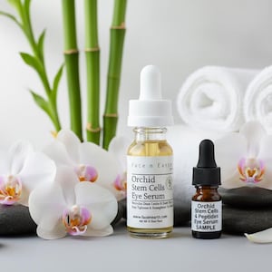 Puede incluir: Dos botellas de Face n Earth Orchid Stem Cells Eye Serum con flores de orqu&iacute;deas blancas, tallos de bamb&uacute; y toallas blancas enrolladas. Las botellas est&aacute;n sobre piedras negras. Una botella tiene un cuentagotas, la otra una muestra.