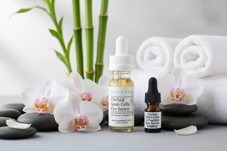Puede incluir: Dos botellas de Face n Earth Orchid Stem Cells Eye Serum con flores de orqu&iacute;deas blancas, tallos de bamb&uacute; y toallas blancas enrolladas. Las botellas est&aacute;n sobre piedras negras. Una botella tiene un cuentagotas, la otra una muestra.