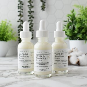 Op de afbeelding: Drie glazen flessen Face n Earth Vit C & B3 Nourishing Serum met witte druppelaars. De flessen zijn gevuld met een romige vloeistof en hebben witte etiketten. Groene planten en witte potten staan op de achtergrond.
