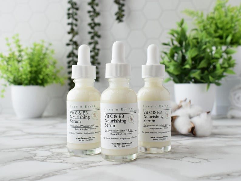 Op de afbeelding: Drie glazen flessen Face n Earth Vit C & B3 Nourishing Serum met witte druppelaars. De flessen zijn gevuld met een romige vloeistof en hebben witte etiketten. Groene planten en witte potten staan op de achtergrond.