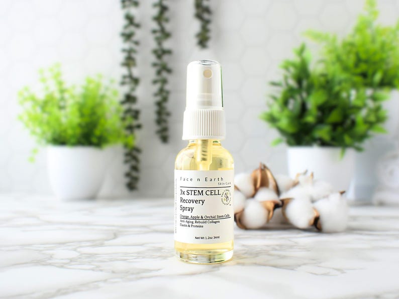 May include: A clear glass bottle with a white spray top and a white label. The label says "3x STEM CELL Recovery Spray" and lists ingredients including orange, apple, and orchid stem cells. The bottle is sitting on a white marble surface.