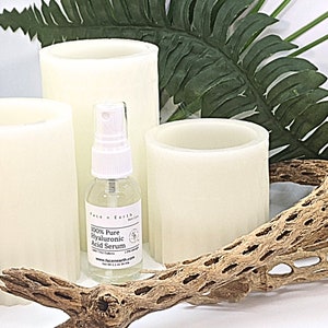 May include: A clear glass spray bottle of Face n Earth 100% Pure Hyaluronic Acid Serum with a white pump top and a label stating 1.1 oz 34 ml, positioned among three off-white cylindrical pillar candles, a green fern leaf, and a piece of cholla wood.
