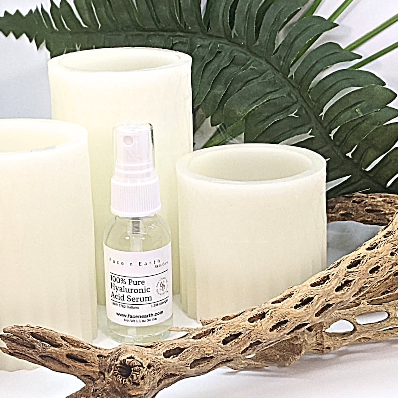 May include: A clear glass spray bottle of Face n Earth 100% Pure Hyaluronic Acid Serum with a white pump top and a label stating 1.1 oz 34 ml, positioned among three off-white cylindrical pillar candles, a green fern leaf, and a piece of cholla wood.