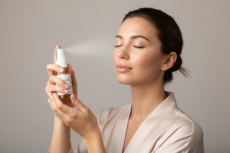 May include: A clear plastic spray bottle with a white pump nozzle and a white label containing black text. The bottle contains a light amber liquid and is emitting a fine mist spray from the nozzle, directed toward the side of the frame.