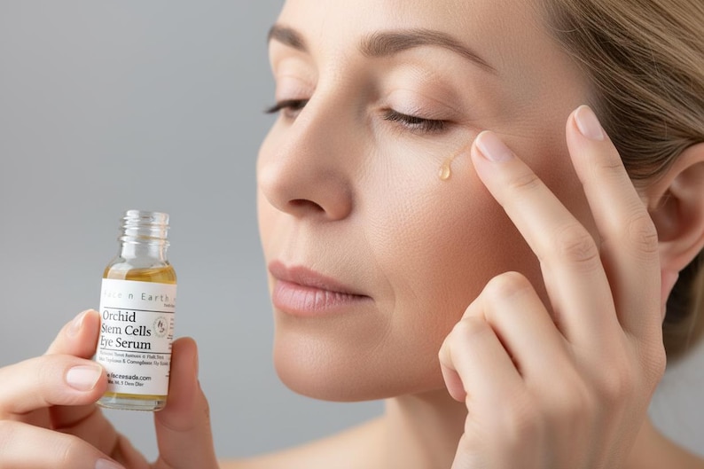 Puede incluir: Una mujer se aplica s&eacute;rum para los ojos. En una mano se sostiene un peque&ntilde;o frasco de vidrio de s&eacute;rum para ojos de c&eacute;lulas madre de orqu&iacute;dea. Se aplica una gota de s&eacute;rum en la piel cerca del ojo. La piel de la mujer es clara.