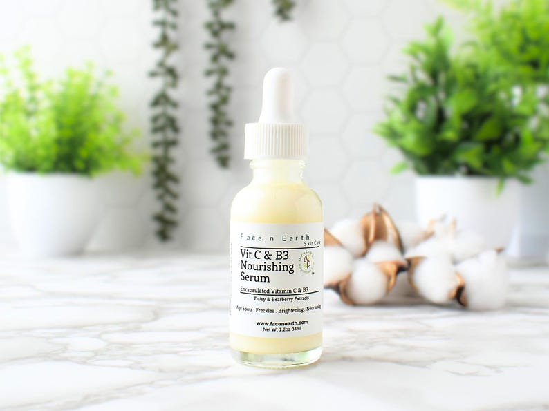 Op de afbeelding: Een heldere glazen fles Face n Earth Vit C & B3 Nourishing Serum met een witte druppelaar. Het etiket bevat tekst over de ingredi&euml;nten en voordelen van het product. De fles is 34 ml. De achtergrond bevat planten en katoen.