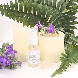 May include: A clear glass spray bottle of Face n Earth 100% Pure Hyaluronic Acid Serum with a white pump top and a label stating Net Wt 1.1 oz. The bottle is surrounded by two cream-colored pillar candles, green fern leaves, purple flowers, and small pink crystals.