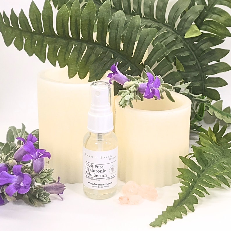 May include: A clear glass spray bottle of Face n Earth 100% Pure Hyaluronic Acid Serum with a white pump top and a label stating Net Wt 1.1 oz. The bottle is surrounded by two cream-colored pillar candles, green fern leaves, purple flowers, and small pink crystals.