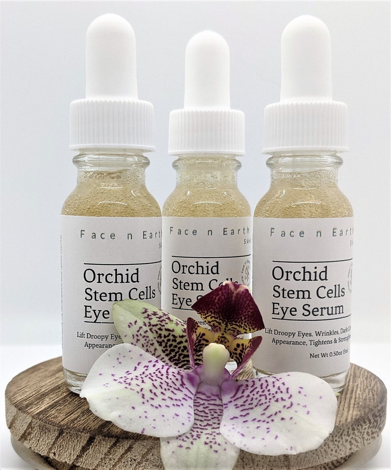 May include: Three glass bottles of orchid stem cell eye serum with white dropper tops. The bottles are labeled "Face n Earth" and "Orchid Stem Cells Eye Serum". The bottles are sitting on a wooden surface with a purple and white orchid flower in front of them.