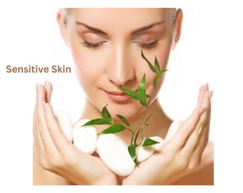 May include: A person with closed eyes holds several smooth white oval stones and a small green bamboo stalk. The text "Sensitive Skin" is printed in brown on the left side of the image, positioned near the person's face.