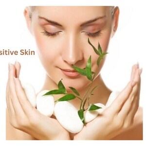 May include: A person with closed eyes holding several smooth white oval stones and a small green bamboo sprig with pointed leaves. The text "Sensitive Skin" is printed in brown letters on the left side of the image against a white background.