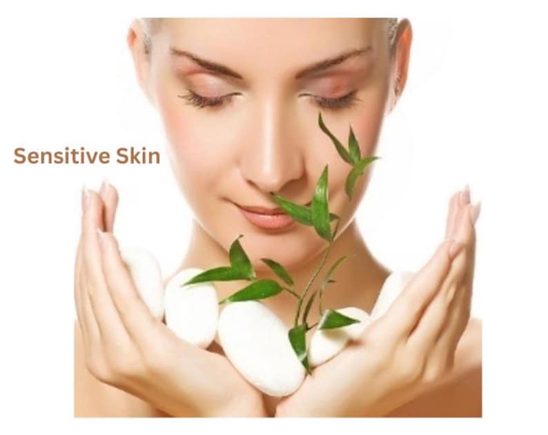 May include: A person with closed eyes holding several smooth white oval stones and a small green bamboo sprig with pointed leaves. The text "Sensitive Skin" is printed in brown letters on the left side of the image against a white background.