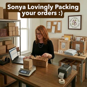May include: A workspace featuring a person in a black long-sleeve shirt packing orders into a cardboard box. The desk includes a digital scale, a laptop, a label printer, and shipping supplies. Text at the top reads: Sonya Lovingly Packing your orders :)