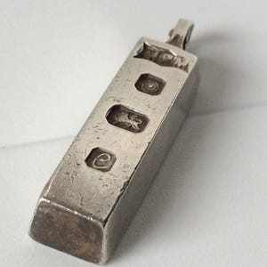 May include: A rectangular silver-coloured metal pendant shaped like a small ingot. The top surface features four distinct stamped hallmarks, including the letters MPM, a circular symbol, a lion figure, and the letter C. It includes a small bail loop.