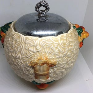May include: A round ceramic jar with a cream-coloured textured surface featuring a raised wheat sheaf design. The jar has a shiny silver-toned metal lid with a decorative handle and is accented with colourful fruit motifs on the sides and base.