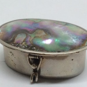 May include: An oval silver trinket box with an iridescent abalone shell lid. The lid features a shimmering display of colours, including green, purple, and blue. The box has a hinged closure and a polished silver finish.