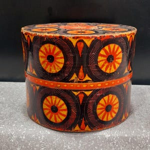 May include: A round, wooden box with a lid, painted with a repeating pattern of orange and yellow circles on a black background. The box is decorated with a band of orange around the middle. The box is sitting on a grey surface.