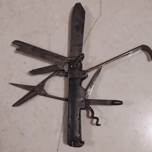 Puede incluir: Una multiherramienta antigua con varios implementos, que incluyen una hoja grande, una sierra, unas tijeras y un sacacorchos. La herramienta está hecha de metal oscuro y muestra signos de edad y uso. Se incluyen herramientas pequeñas adicionales.