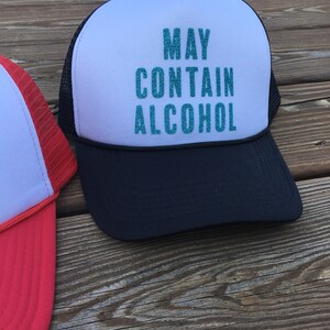 Może przedstawiać: Czarno-biała czapka trucker z białym panelem z przodu, na kt&oacute;rym napis "MAY CONTAIN ALCOHOL" w turkusowych literach z brokatem.