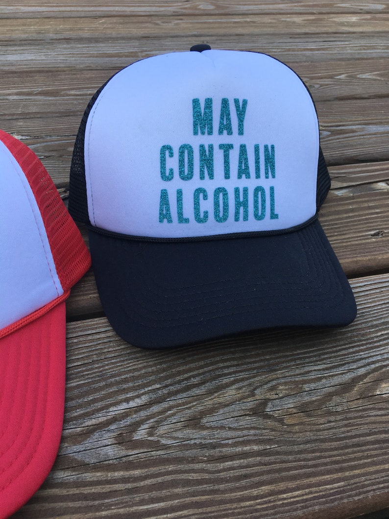 Może przedstawiać: Czarno-biała czapka trucker z białym panelem z przodu, na kt&oacute;rym napis "MAY CONTAIN ALCOHOL" w turkusowych literach z brokatem.