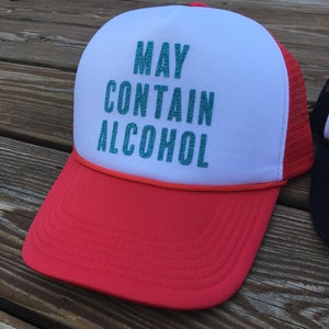 Może przedstawiać: Czapka truckerowa w kolorze czerwonym i białym z siateczką z tyłu. Z przodu czapki znajduje się napis "MAY CONTAIN ALCOHOL" w turkusowym brokacie.