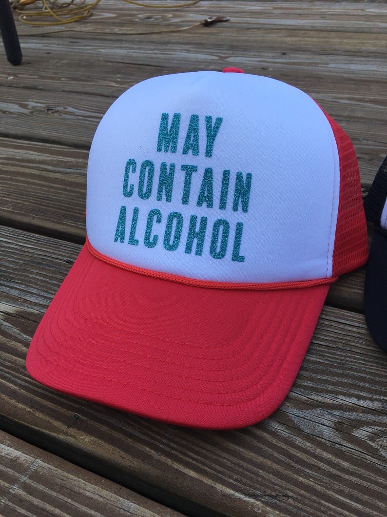 Może przedstawiać: Czapka truckerowa w kolorze czerwonym i białym z siateczką z tyłu. Z przodu czapki znajduje się napis "MAY CONTAIN ALCOHOL" w turkusowym brokacie.