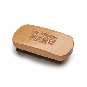 Könnte beinhalten: Eine Holz-Bartbürste mit schwarzen Borsten. Die Bürste trägt die Gravur "The Bearded Man Co." auf der Oberseite.