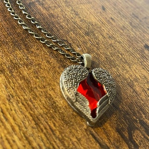 May include: A gold chain necklace with a heart-shaped pendant. The pendant features a red gemstone surrounded by detailed metal wings.