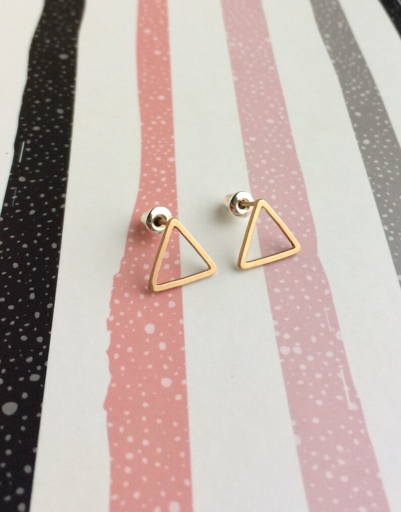 Puede incluir: Un par de pendientes de tri&aacute;ngulo dorado con un dise&ntilde;o simple y minimalista. Los pendientes est&aacute;n sobre un fondo a rayas.