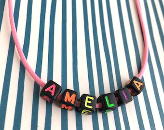 Puede incluir: Un collar de cordón rosa con cuentas cuadradas negras que deletrean "AMELIA" en letras coloridas. Las cuentas tienen una fuente vibrante y multicolor. El fondo es un patrón de rayas azules y blancas.