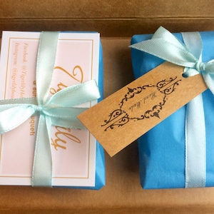 Puede incluir: Dos peque&ntilde;os regalos envueltos en papel de seda azul con cinta azul claro y una etiqueta marr&oacute;n que dice "Hecho a mano". Un regalo es una tarjeta con fondo rosa y el texto "Realmente un hermoso ramo" y el otro regalo es una peque&ntilde;a caja.